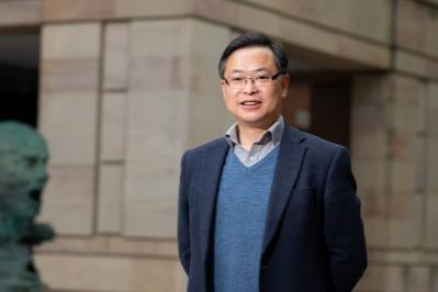 Chinese male member of staff with short dark hair, wearing glasses, a navy blazer over a grey shirt and blue v neck jumper, standing in front of the Maclay library, A beige stone building with a head sculpture to his right